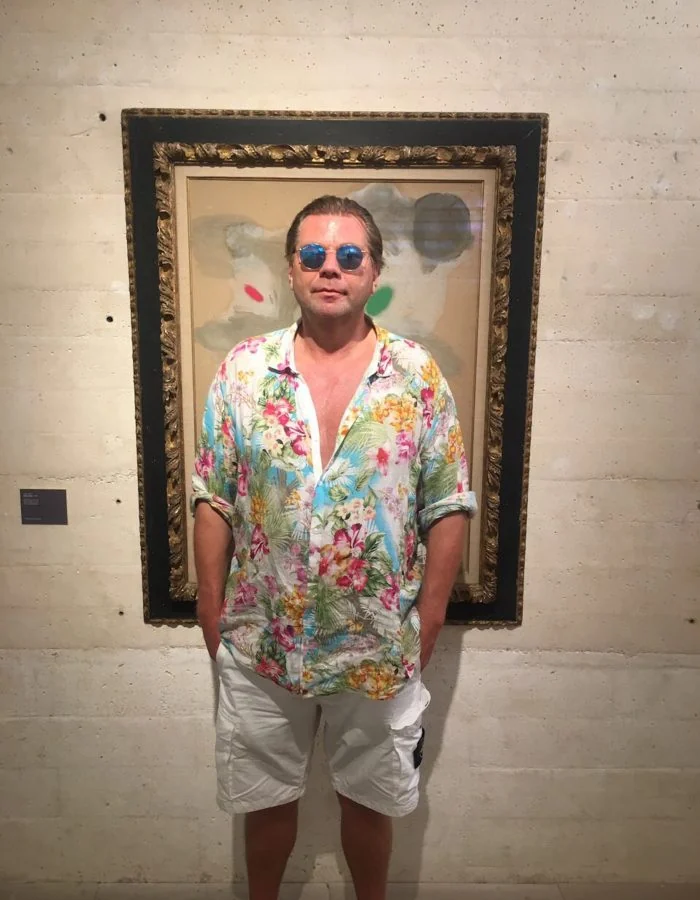 Markus Tollmann in a floral shirt standing in front of a large framed work on a concrete wall